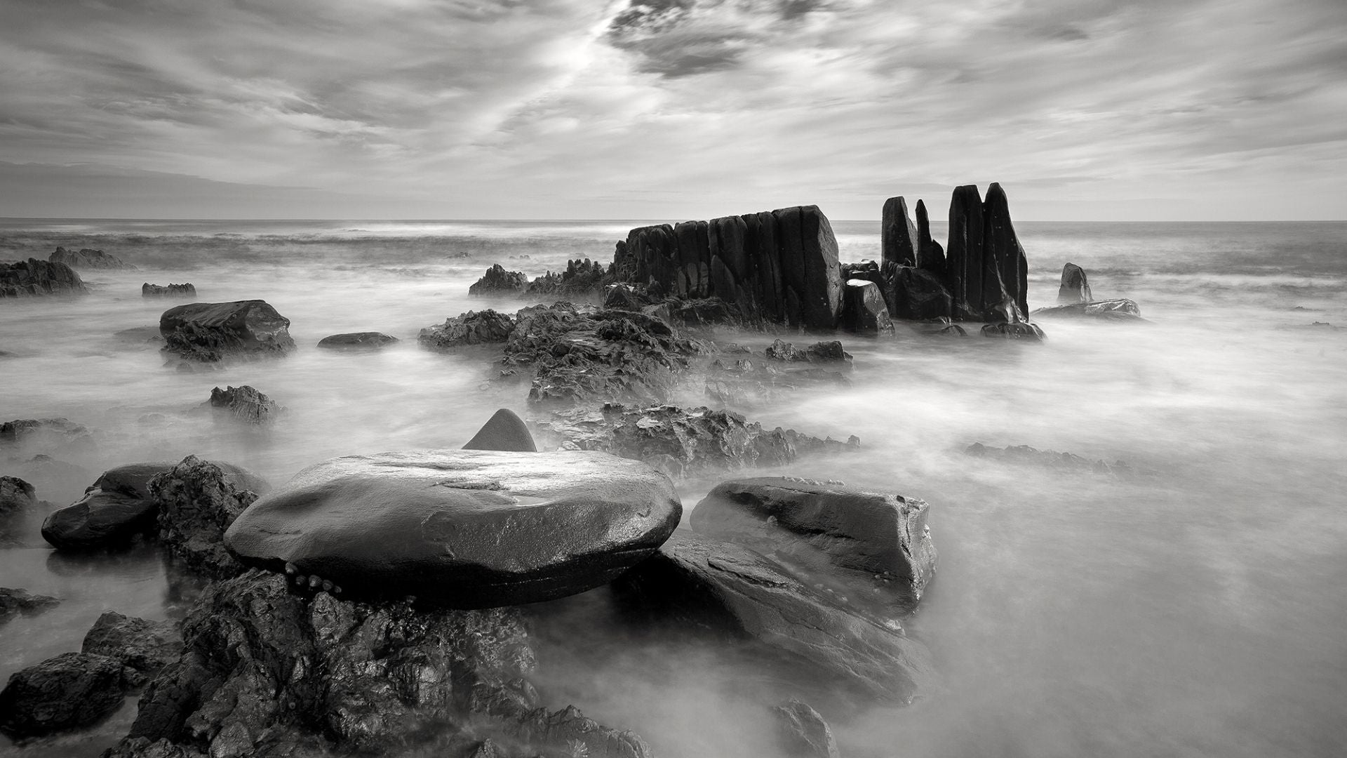 ChatGPT said:  Black and white seascape capturing rugged rocks along the shoreline, with misty waves rolling under a dramatic sky.