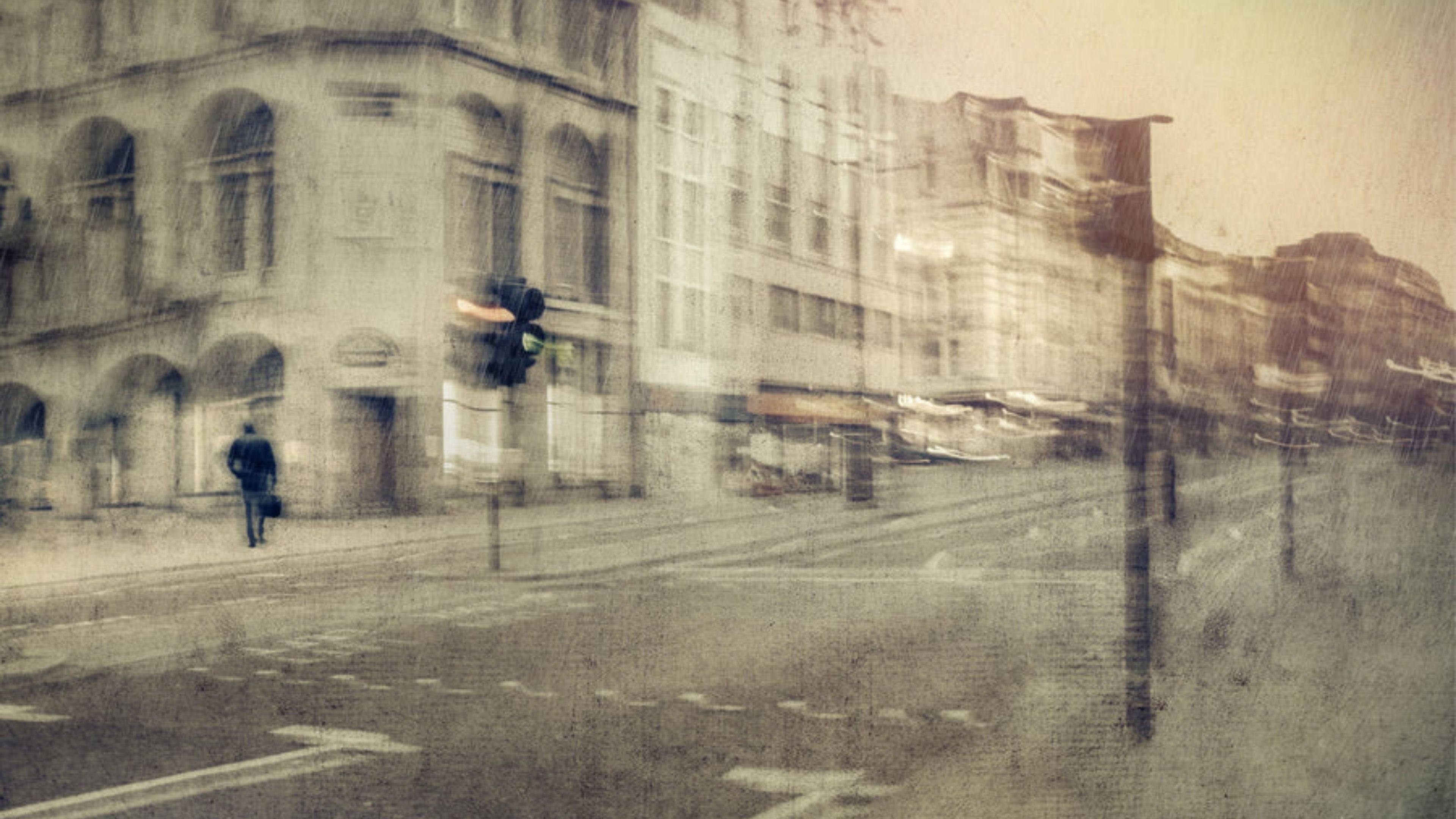 Abstract urban street scene with blurred buildings, a lone pedestrian, and a traffic light in soft muted tones, conveying a moody city atmosphere.