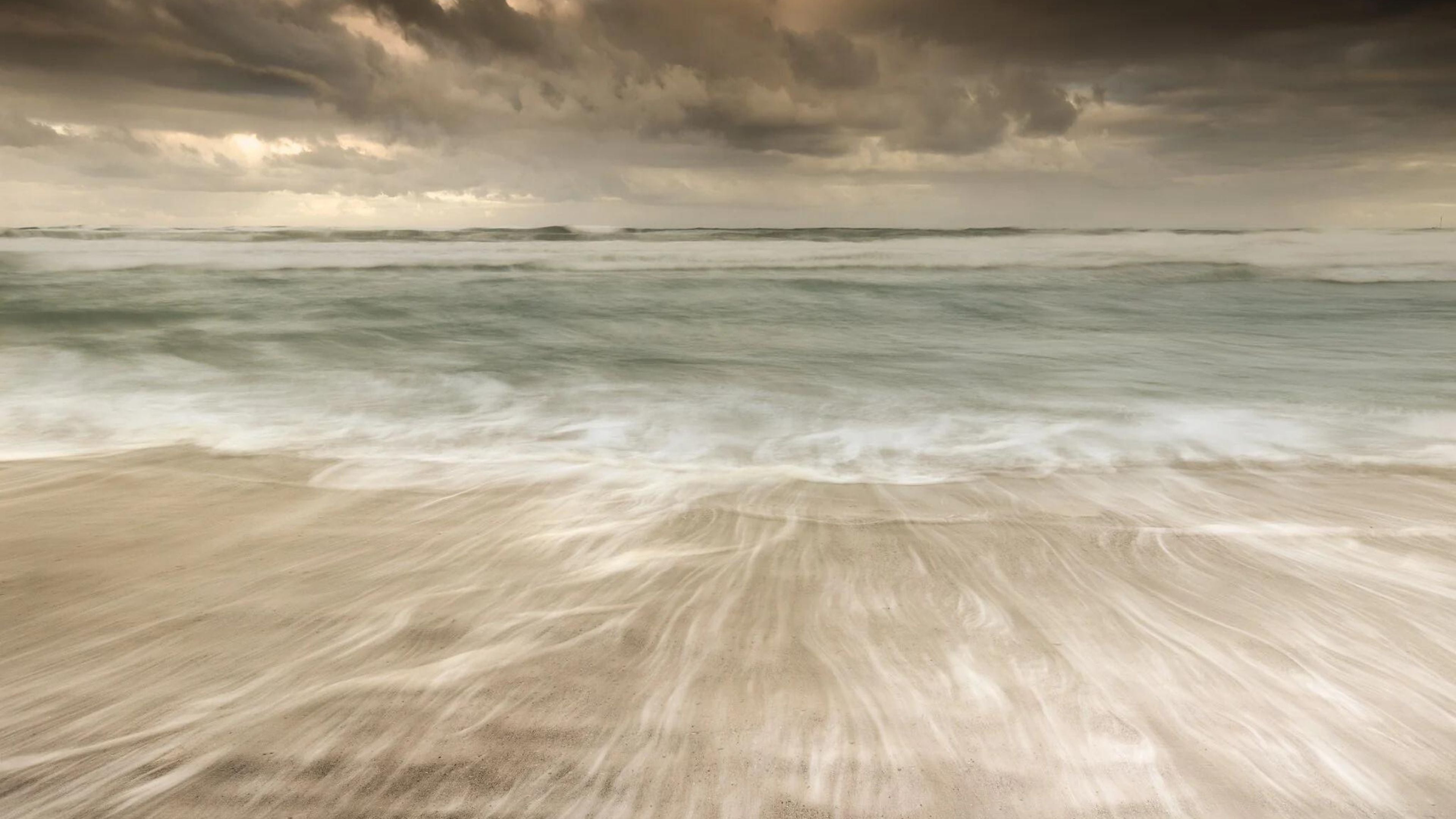 Fine art seascape with sweeping sand patterns and soft waves under dramatic cloudy skies, captured in long-exposure coastal photography