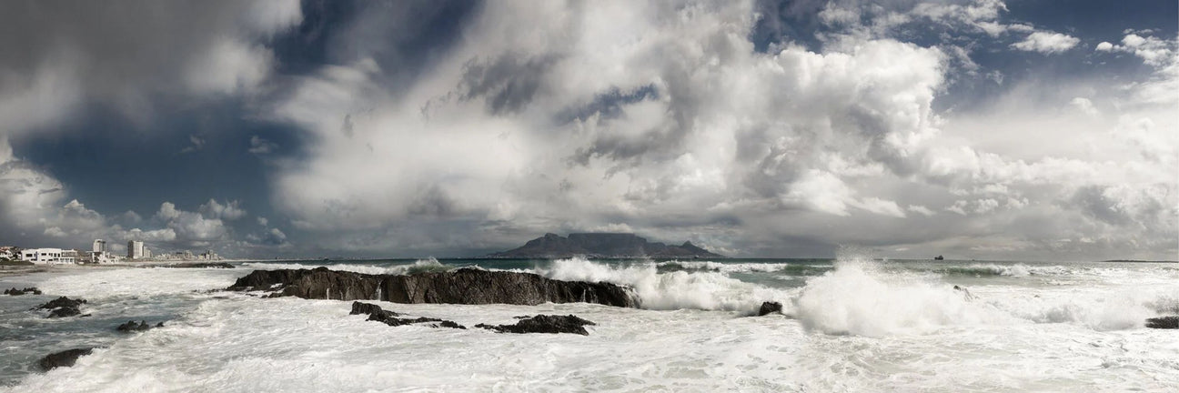 Panoramic fine art photograph of Table Mountain viewed from the Blouberg Strand coast, with dramatic waves crashing against rocks under a stormy Cape Town sky.